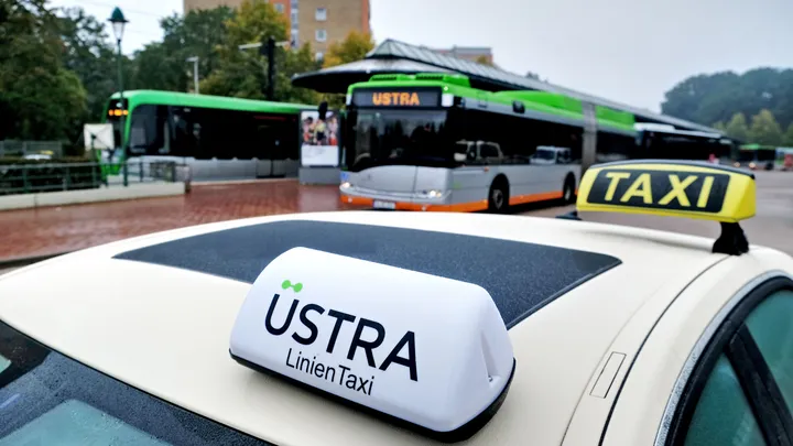 Dach eines ÜSTRA Linien Taxis an Haltestelle. Ein Bus und eine Bahn sind im Hintergrund zu erkennen.