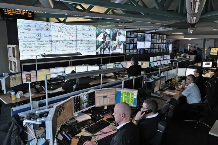 Mitarbeitende sitzen in zwei Reihen an Computerarbeitsplätzen mit fünf Bildschirmen. An der Wand vor ihnen sind große Monitore. Dort sieht man eine schematische Darstellung der Stadtbahnlinien und Bilder der Überwachungskameras an Haltestellen.