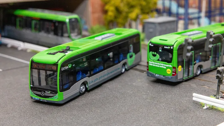 Zwei Modellbusse eCitaro fahren in einer Modellbaulandschaft in jeweils eine andere Richtung. Im Hintergrund sieht man eine Ampel, Schienen, einen TW 3000, einen Tunnel, Gebäude und Bäume.