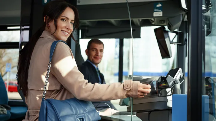 Ein Fahrgast steht vorne im Bus und hält eine Bankkarte an das Lesegerät. Im Hintergrund sitzt ein Fahrpersonal. Beide Menschen lächeln.