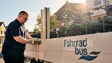 On the left side of the picture, a man wearing glasses and work gloves is operating the latch on the bicycle trailer. The trailer is white and has an open loading area with several bicycles on it. On the right side of the picture, the blue words “Fahrradbus” (bicycle bus) can be seen on the trailer.