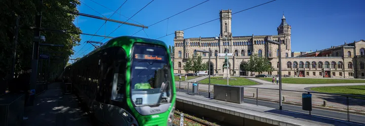 Links im Bild steht die ÜSTRA Straßenbahn, Linie 5 in Richtung Anderten, an der Haltestelle "Leibniz Universität". Im Hintergrund sind Gleise, die Universität, Menschen und Bäume zu sehen.