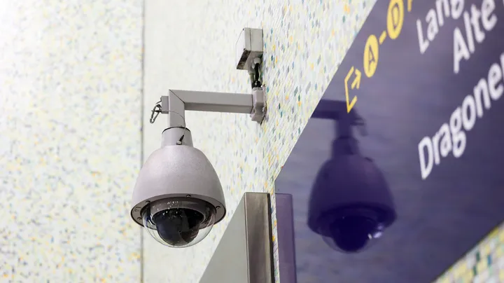 Eine Überwachungskamera, die an der Wand einer Stadtbahnstation befestigt ist. Im Hintergrund ist ein blaues Navigationsschild zu erkennen.