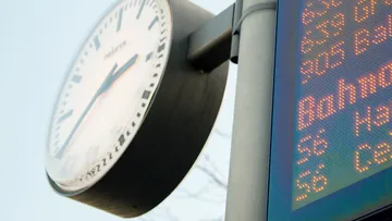 Nahaufnahme einer Bahnhofsuhr vor blauem Himmel, im Anschnitt ist eine digitale Anzeigentafel zu erkennen