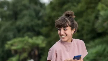 Eine junge Frau sitzt im Schneidersitz am Maschsee und lacht, im Hintergrund ist ein Gebäudekomplex sichtbar