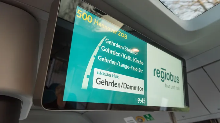 Ein zweigeteilter Bildschirm hängt oben im Innenraum eines Busses. Auf dem Bildschirm sieht man die nächsten Haltestellen der Linie 500 in Gehrden: Dammtor, Lange-Feld-Straße, Katholische Kirche, Steintor. Auf der zweiten Hälfte des Bildschirms steht das regiobus Logo.
