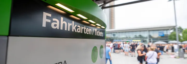 Links im Bild ist der obere Teil von einem ÜSTRA Fahrkartenautomat zu sehen. Im Hintergrund sind viele Menschen zu sehen, die verschwommen sind.