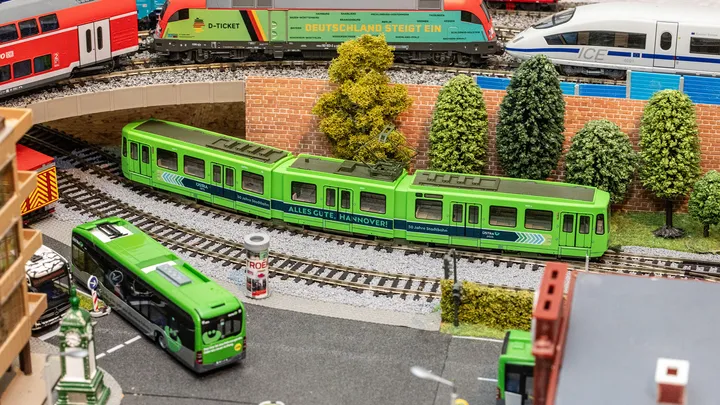 Der TW 6001 fährt in einer Modellbaulandschaft auf Schienen. Auf seiner Seite ist eine Beklebung: "Alles Gute Hannover". Vor der Bahn sthet ein eCitaro Modell. Dort sind ebenfalls Straßenlaternen, Gebäude, eine Uhr und eine Litfaßsäule. Im Hintergrund sind einige Bäume und Nahverkehrszüge.