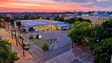 Luftaufnahme der Betriebswerkstatt Döhren im Morgengrauen. Neben der Halle steht ein TW 3000. Um das Werkstattsgebäude herum stehen viele grüne Bäume.