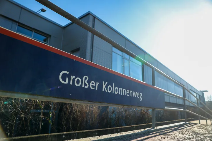 Zu sehen ist ein Haltestellenschild auf dem "Großer Kolennenweg" steht. Die Schrift ist weiß, der Hintergrund des Schildes dunkelblau. Im Hintergrund sieht man ein Gebäde mit Flachdach, blauen Himmel und die Sonne.