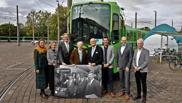 Ein TW 6000 mit der Liniennummer 12 und dem Ziel "Hauptbahnhof" steht auf dem Betriebshof. Vor der Bahn stehen viele Menschen, die ein großes Bild halten. Das Bild zeigt die Eröffnung der Stadtbahn vor 50 Jahren. Es ist schwarz-weiß. Auf dem Bild ist ein TW 6000 in einer Tunnelstation mit der Liniennummer 12 und dem Ziel "Hannover. Vor der Bahn stehen viele Leute. Einige winken.
