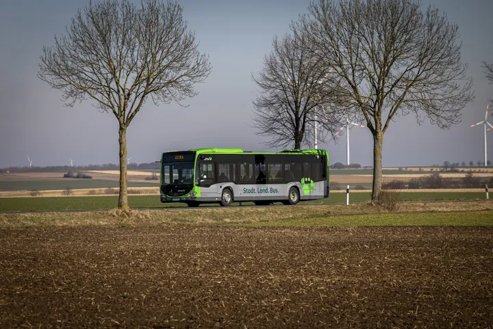 Ein ÜSTRA Bus fährt auf einer Landstraße zwischen Feldern. Auf dem Bus ist das ÜMO Logo zu sehen und die Aufschrift "Stadt. Land. Bus". Im Hintergrund sind Felder, Bäume und Windräder zu sehen.
