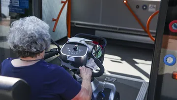 Man sieht eine alte Frau von hinten mit grauen Haaren, die in einem elektrischen Rollstuhl sitzt und damit gerade durch die geöffneten Türen in einen Bus einsteigt.