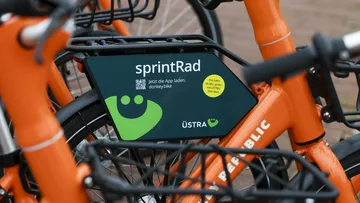 Nahaufnahme auf orangefarbige Sprinträder. Im Fokus des Bildes ist ein dunkelgrünes Schild, das an einem Gepäckträger montiert ist mit der Aufschrift "sprintRad" und dem hellgrünen ÜMO.