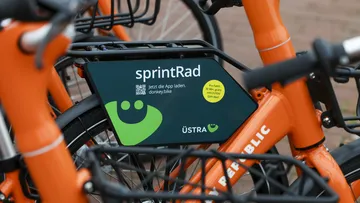 Nahaufnahme auf orangefarbige Sprinträder. Im Fokus des Bildes ist ein dunkelgrünes Schild, das an einem Gepäckträger montiert ist mit der Aufschrift "sprintRad" und dem hellgrünen ÜMO.