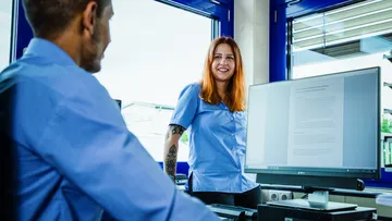 Ein Azubi sitzt am PC und lächelt zu einer Azubi, die hinter dem PC am Fenster steht