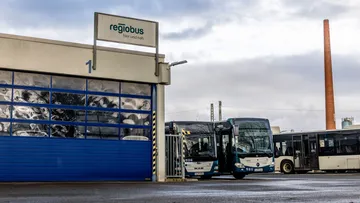 Links ist das blaue Rolltor der Bushalle zu sehen. Am Dach ist ein Schild mit dem Logo der regiobus GmbH angebracht. Rechts neben der Halle stelehn zwei Hybridbusse. Im Hintergrund steht ein Industrieschornstein.