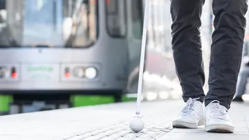 Nahaufnahme auf ein Blindenleitsystem an einer Stadtbahnstation. Zu sehen sind ein Blindenstock, Füße und im Hintergrund eine Bahn.