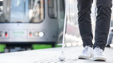 Nahaufnahme auf ein Blindenleitsystem an einer Stadtbahnstation. Zu sehen sind ein Blindenstock, Füße und im Hintergrund eine Bahn.