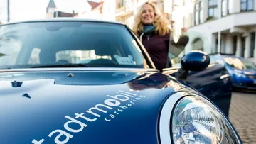 Zu sehen ist ein Mini Cooper von vorne, im Fokus ein Scheinwerfer und eine stadtmobil-Beklebung. Eine blonde Frau steht hinter der geöffneten Fahrertür.