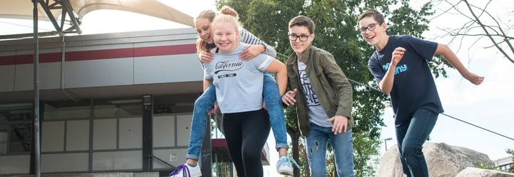 Zwei Mädchen und zwei Jungs laufen lachend in Richtung der Kamera. Ein Mädchen hat das andere Mädchen Huckepack, die Jungs tragen Brillen.