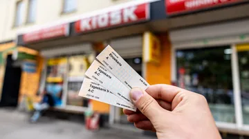 Eine Hand hält drei ÜSTRA Fahrkarten in die Kamera. Im Hintergrund ist ein Kiosk zu sehen und eine Person sitzt links vor dem Kiosk auf einer Bank.