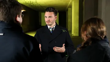 Ein Mann in ÜSTRA Dienstkleidung steht in einem Tunnel und erzählt zwei Menschen etwas, die von hinten zu sehen sind.