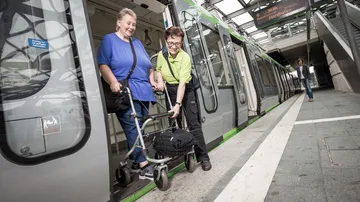 Eine Person mit Rollator verlässt mit Hilfe des Fahrgastbegleitservice eine Stadtbahn, die in einer überdachten Stadtbahnstation steht.