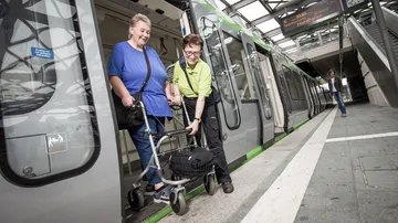 Eine Person mit Rollator verlässt mit Hilfe des Fahrgastbegleitservice eine Stadtbahn, die in einer überdachten Stadtbahnstation steht.