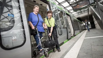 Eine Person mit Rollator verlässt mit Hilfe des Fahrgastbegleitservice eine Stadtbahn, die in einer überdachten Stadtbahnstation steht.
