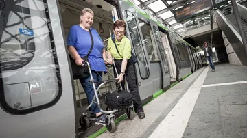 Eine Person mit Rollator verlässt mit Hilfe des Fahrgastbegleitservice eine Stadtbahn, die in einer überdachten Stadtbahnstation steht.