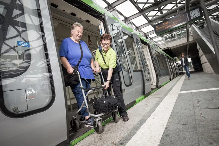 Eine Person mit Rollator verlässt mit Hilfe des Fahrgastbegleitservice eine Stadtbahn, die in einer überdachten Stadtbahnstation steht.