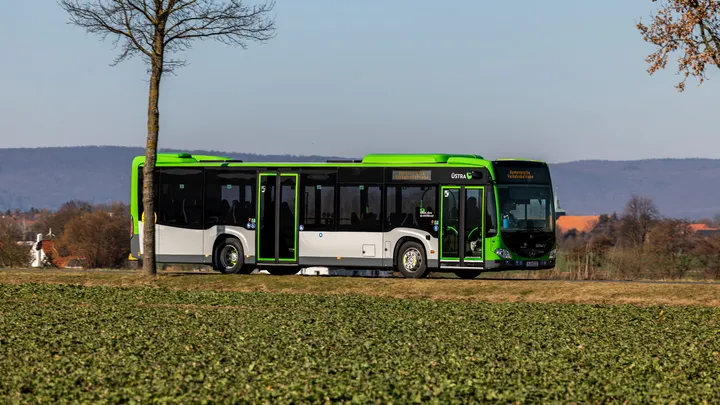 Ein Citaro Bus mit Hybrid Antrieb fährt auf einer Landstraße. Im Vordergrund ist ein grünes Feld und zwei Bäume am Straßenrand.