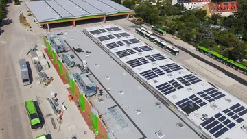 Eine Luftaufnahme des Betriebshofes Mittelfeld. Man sieht zwei Bushallen hintereinander. Das Dach der vorderen Halle hat eine Solaranlage. Links neben der vorderen Halle stehen drei Busse.