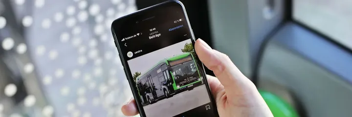 Eine Hand hält ein Handy. Das WLAN-Symbol ist aktiviert. Auf dem Bildschirm sieht man den Instagramkanal der ÜSTRA mit einem Foto eines eCitaro Busses. Im Hintergrund sieht man verschwommen das Sitzpolster und Fenster eines Busses.