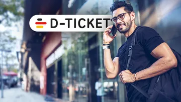Männlicher, telefonierender Fußgänger in der Innenstadt vor einer Ladenzeile und Signet des Deutschlandtickets