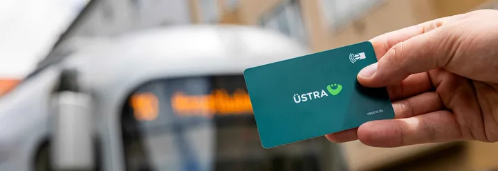 Im Bild wird eine Chipkarte des Deutschlandttickets hochgehalten. Im Hintergrund steht eine Stadtbahn, der TW 2000