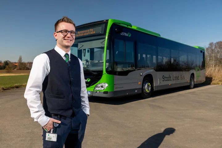 Ein Mitarbeiter der regiobus Hannover GmbH steht vor einem ÜSTRA Bus und lächelt in die Kamera. Im Hintergrund sind der Bus, Felder, Bäume und Dächer zu sehen.