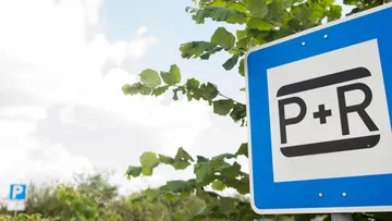 Rechts im Bild ist ein blau-weißes Schild mit der Aufschrift "P+R" zu sehen, im Hintergrund grüne Blätter eines Baumes. Links im Bild sind blauer Himmel und weiße Wolken zu sehen.
