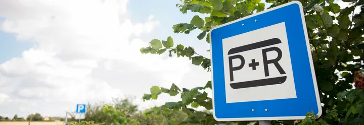 Rechts im Bild ist ein blau-weißes Schild mit der Aufschrift "P+R" zu sehen, im Hintergrund grüne Blätter eines Baumes. Links im Bild sind blauer Himmel und weiße Wolken zu sehen.