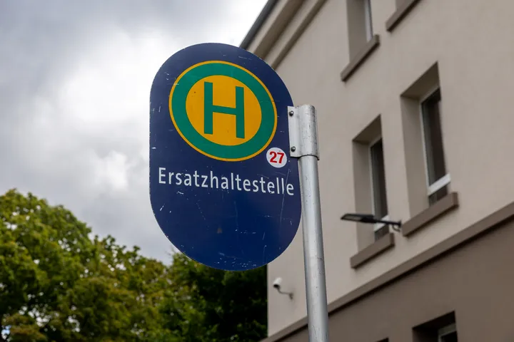 Im Fokus des Bildes ist das Schild einer Haltestelle zu sehen. Das Schild ist blau. Ein grünes großes H auf einem gelben Kreis. Darunter steht Ersatzhaltestelle. Im Hintergrund sieht man ein Stück Himmel mit grauen Wolken, rechts eine Baumkrone, links die Fassade eines Hauses.