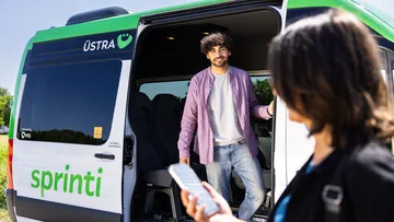 Ein Mann steht in der geöffneten Tür eines ÜSTRA sprinti Fahrzeugs. Vor dem Fahrzeug wartet eine Frau mit einem Handy in der Hand.