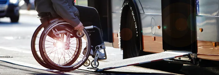 Eine Person im Rollstuhl, die über die ausgefahrene Rampe in einen Bus fährt. Im Fokus des Bildes sind Rollstuhl und Rampe, Person und Bus sind nur abgeschnitten zu sehen.