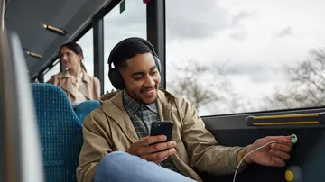Im Bus sitzt ein Fahrgast mit Kopfhörern und einem Smartphone in der Hand. Er steckt ein U S B Ladekabel in einen Ladeanschluss in der Wand des Busses.