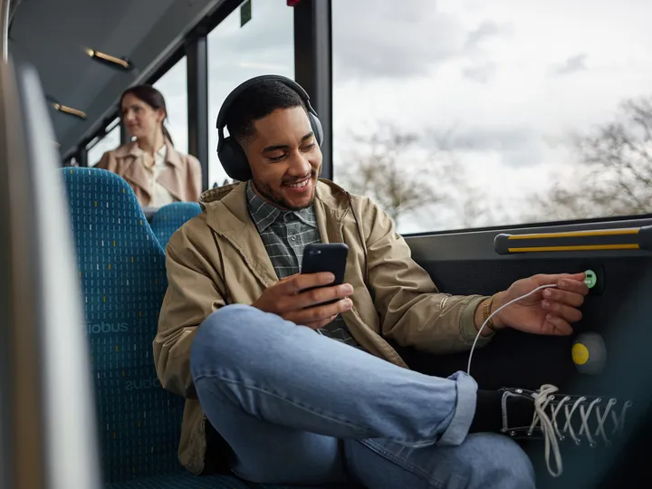 Im Bus sitzt ein Fahrgast mit Kopfhörern und einem Smartphone in der Hand. Er steckt ein U S B Ladekabel in einen Ladeanschluss in der Wand des Busses.