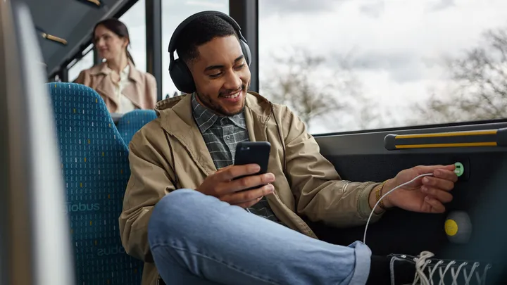 Im Bus sitzt ein Fahrgast mit Kopfhörern und einem Smartphone in der Hand. Er steckt ein U S B Ladekabel in einen Ladeanschluss in der Wand des Busses.