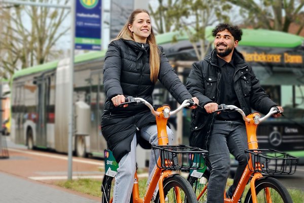 Coverbild des GVH journals 01.2024: Zwei Personen fahren jeweils auf einem orangenen Fahrrad an einer Bushaltestelle vorbei.