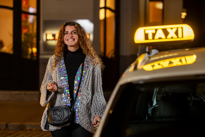 Eine Frau steht neben einem Taxi und schaut lächelnd in die Kamera. Das Taxi-Symbol leuchtet, im Hintergrund sieht man Fenster eines Gebäudes, die ebenfalls beleuchtet sind.
