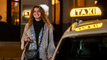 Eine Frau steht neben einem Taxi und schaut lächelnd in die Kamera. Das Taxi-Symbol leuchtet, im Hintergrund sieht man Fenster eines Gebäudes, die ebenfalls beleuchtet sind.