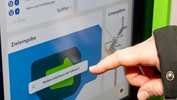 Eine Hand ist zu sehen, der Zeigefinger zeigt auf die Zieleingabe am Fahrkartenautomat der ÜSTRA. Hier steht: "Wohin möchtest du fahren?".